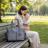Woman holding a baby and sitting on a bench with a large gray diaper bag in a park.